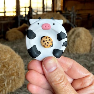 May include: A whimsical, handmade cow-shaped dish with black spots and a pink nose, holding a miniature chocolate chip cookie. The dish is white and the cookie is brown with chocolate chips. The design is playful and charming.