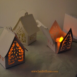 Peut inclure: Trois lanternes blanches en forme de maison en papier, avec des motifs découpés complexes. L'une est ouverte, révélant une bougie allumée. Les deux autres sont fermées. Les lanternes présentent un motif de sapin de Noël et sont éclairées de l'intérieur.