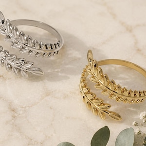 May include: Two rings, one silver and one gold, each with a leaf design that wraps around the finger. The rings are displayed on a light, marble-effect surface, with small white flowers and greenery in the background.