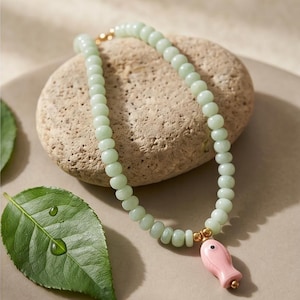Puede incluir: Un collar que presenta una hilera de cuentas lisas de color verde pálido y un colgante individual con forma de pez rosa. El colgante está decorado con pequeñas cuentas de metal dorado a ambos lados y el collar se cierra con un broche dorado.