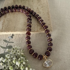 Op de afbeelding: Een ketting met ronde, donkerpaarse kralen afgewisseld met kleine, platte goudkleurige tussenstukjes. De ketting bevat een grote, heldere, ovaalvormige gefacetteerde hanger in een goudkleurige zetting aan de onderkant van de kralenstreng.