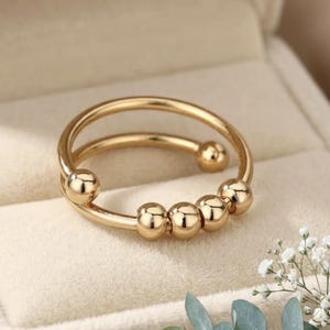 May include: A gold-coloured ring with a double band design. The ring features five small, spherical beads that can be moved along the lower band. The ring is displayed on a soft, cream-coloured surface.