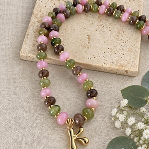 Puede incluir: Un collar de cuentas con cuentas redondas alternas de color rosa, verde oliva y marrón oscuro separadas por pequeños espaciadores dorados. El collar incluye un colgante con la letra K dorada que cuelga del centro de la cadena.