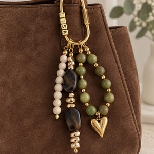 May include: A gold-toned carabiner keyring attached to a brown suede handbag. The keyring features the name LISA in block letters, a strand of cream beads, a strand of dark brown marbled beads, and a loop of green beads with a gold heart charm.