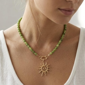 May include: A necklace featuring a strand of small, round, olive green beads interspersed with tiny gold-toned spacers. The necklace includes a central gold-toned sun pendant with a spiral centre and radiating points, attached by a gold-toned link.