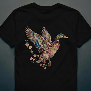 May include: Black t-shirt featuring a colorful floral duck design with outstretched wings. The duck's body and wings are adorned with various flowers and patterns. A trail of small flowers follows the duck.