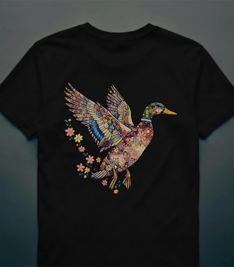 May include: Black t-shirt featuring a colorful floral duck design with outstretched wings. The duck's body and wings are adorned with various flowers and patterns. A trail of small flowers follows the duck.
