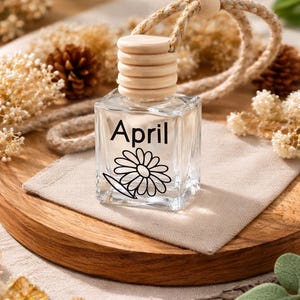 May include: A clear, square glass air freshener bottle with a wooden cap and a woven rope. The bottle has the word "April" and a daisy illustration. It sits on a beige linen cloth on a wooden tray.
