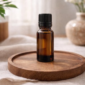 May include: A small amber glass bottle with a black cap sits on a round wooden tray. The bottle is filled with a clear liquid. The tray is on a beige surface, with a blurred background of plants and a vase.
