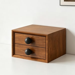 May include: A small wooden storage unit with a rich brown wood grain finish, featuring two stacked drawers. Each drawer is equipped with a single, large, matte black spherical knob centered on the front panel of the drawer face.