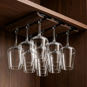 May include: A dark wood wine glass rack mounted under a cabinet, holding six clear glass wine glasses hanging upside down by their bases. The rack features parallel slots for sliding the stems of the glasses into place for organized storage.