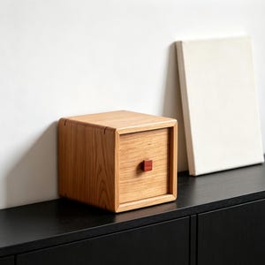 May include: A square wooden storage box with a light brown grain finish and a single drawer. The drawer features a small, dark red square knob centered on the front panel. The box has rounded edges and a smooth, polished surface texture.