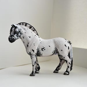 May include: A white horse figurine with black spots and a black mane. The horse is standing in a profile position, showing a textured coat, dark hooves, and a detailed face with black markings around the muzzle and eyes, captured in a static pose.
