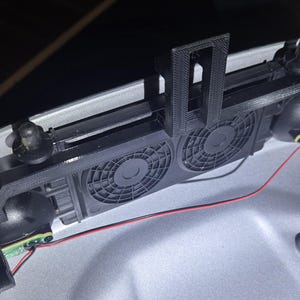 May include: Black 3D-printed device with two circular fans, a bracket, and a red wire. The device is mounted on a light gray surface. The fans have a radial design.