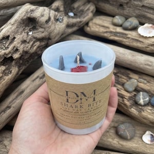 May include: A soy wax candle in a clear glass jar with a tan paper label reading DM Dorset Manor Shark Bite. The light blue wax features three grey shark fin shapes and a red wax accent near the central wick, indicating a 5 oz net weight.