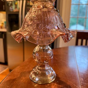 Pode incluir: Uma lâmpada vintage de vidro rosa com uma sombra com babados e detalhes florais. A lâmpada tem uma base redonda e uma haste decorativa. A sombra é adornada com folhas e flores em relevo. A lâmpada está em uma mesa de madeira.