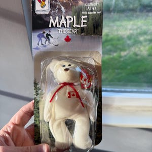 May include: A white plush bear toy named "Maple" with a red ribbon and a Canadian flag emblem. The bear is packaged in a clear plastic casing with a cardboard backing featuring a mountain scene and the text "Collect All 4!".