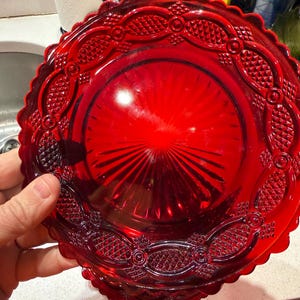 Könnte beinhalten: Ein leuchtend roter Glasteller mit dekorativem, gewelltem Rand. Der Teller hat ein aufwendiges geprägtes Muster am Rand und ein radiales Design in der Mitte. Der Teller wird von einer Hand gehalten.
