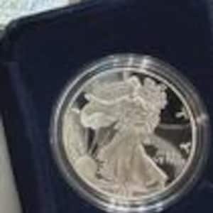 Puede incluir: Una moneda de plata con una representación detallada de una figura caminando, posiblemente un ángel o una diosa, con el sol saliendo al fondo. La moneda está dentro de un soporte de plástico transparente, sobre un fondo de terciopelo azul oscuro.