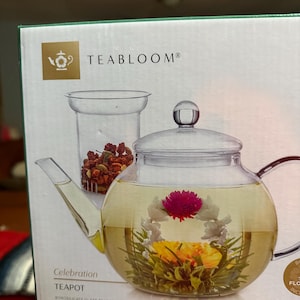May include: A clear glass teapot with a built-in infuser and a blooming tea flower inside. The teapot is labeled "TEABLOOM" and "Celebration TEAPOT". The infuser contains loose tea leaves. The teapot is designed for brewing tea.