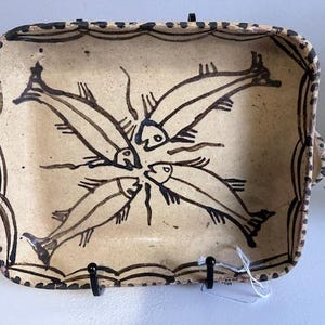 May include: A rectangular ceramic dish with a beige background and a black fish design. The dish has a scalloped edge and two small handles. The fish are arranged in a circular pattern in the center of the dish.