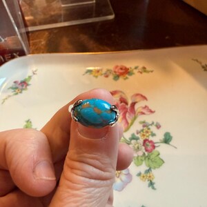 May include: A silver ring featuring a large, oval turquoise stone with brown veining. The ring is held up against a floral patterned background, showcasing the intricate details of the jewelry.