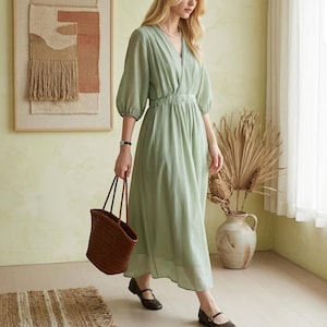 May include: A sage green midi dress with a V-neckline, three-quarter length puff sleeves, and an elasticated waist. The dress features a soft, textured fabric and a flowing skirt. A dark brown woven leather tote bag is carried alongside the dress.