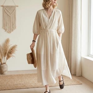May include: A cream-coloured midi dress featuring a V-neckline with delicate pearl-like trim, three-quarter length sleeves, and an elasticated waist. The dress has a flowing skirt with side slits and is paired with a woven straw hat.