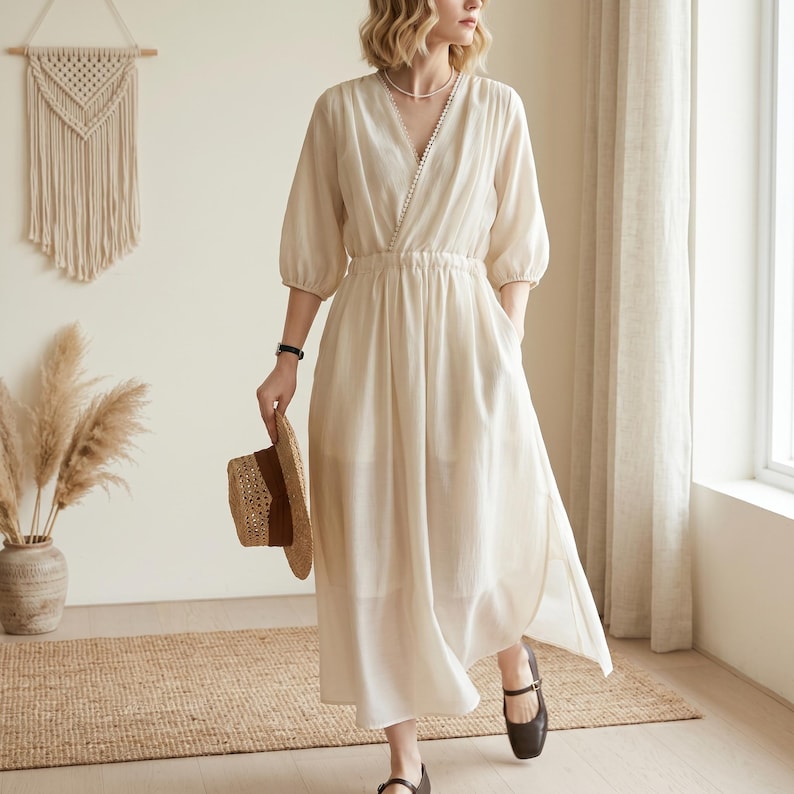 May include: A cream-coloured midi dress featuring a V-neckline with delicate pearl-like trim, three-quarter length sleeves, and an elasticated waist. The dress has a flowing skirt with side slits and is paired with a woven straw hat.