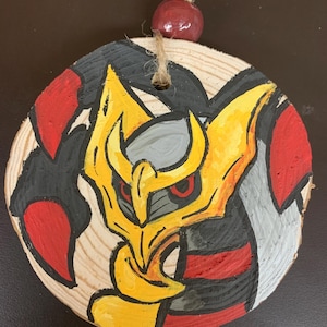 May include: Hand-painted wooden ornament featuring a yellow, red, gray, and black character design. The ornament is round, made of a wood slice, and has a jute string and a red bead for hanging. The design is centered on the wood.