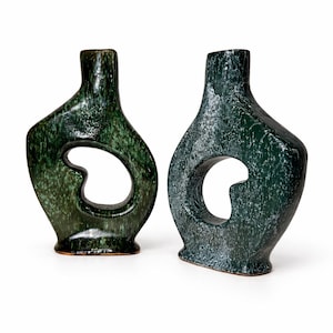 Peut inclure: Deux vases en céramique vert foncé de forme organique unique. Chaque vase présente une grande ouverture irrégulière au centre. Les vases ont une surface texturée et une finition brillante, ajoutant de l'intérêt visuel.