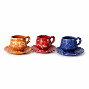 Peut inclure: Trois tasses à café et soucoupes en céramique orange, rouge et bleu. Chaque tasse et soucoupe présente un motif blanc moucheté. Les tasses ont des corps arrondis et de petites anses. Les soucoupes sont rondes et plates. L'ensemble est sur fond blanc.