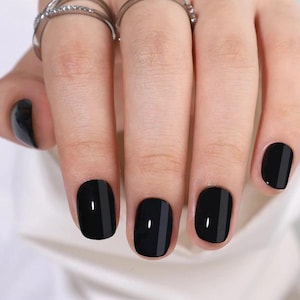 Puede incluir: Primer plano de una mano con uñas negras pintadas. Cada uña está pintada de negro con una raya gris vertical en el centro. La mano también tiene varios anillos plateados.