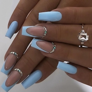 Op de afbeelding: Lichtblauwe vierkante acrylnagels met een French tip design. De nagels hebben een nude basis met een lichtblauwe omtrek en een rij kleine, sprankelende strass steentjes. Een zilveren ring met een grote, vierkant geslepen steen is ook zichtbaar.