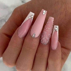 May include: A hand displaying long, coffin-shaped acrylic nails. The nails feature a variety of designs, including a matte pink, a glitter-covered nail, and nails with white tips and rhinestone accents. The nails are a light pink color.