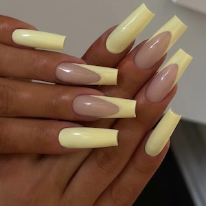 May include: A hand displaying long, square-shaped artificial nails. The nails feature a pale yellow tip and a nude base. The manicure presents a contemporary twist on the traditional French tip design.