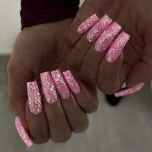 May include: Pink glitter acrylic nails. The nails are long, square-shaped, and covered in a sparkling pink glitter. The nails are on a hand with a burgundy sleeve in the background.