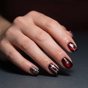 May include: A hand with manicured nails. The nails are painted a deep red with silver designs. Some nails feature a swirl, while others have silver accents. The overall design is elegant and eye-catching.