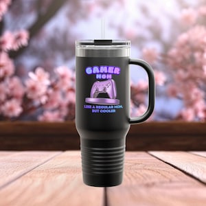 Puede incluir: Taza de viaje negra con asa y pajita. La taza presenta el texto "GAMER MOM" en letras de neón moradas, junto con un gráfico de mando de juego morado. También se muestra el texto "LIKE A REGULAR MOM, BUT COOLER".