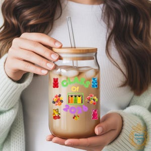 May include: A clear glass tumbler with a bamboo lid and a straw, filled with a light-coloured beverage and ice. The tumbler is decorated with colourful gummy bear stickers and the text "CLASS OF 2026".