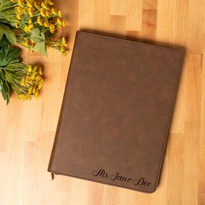 May include: A brown leather-bound journal with the inscription "Ms. Jane Doe" in elegant script. The journal lies on a wooden surface, with yellow flowers and green leaves in the upper left corner. The journal is a classic gift.