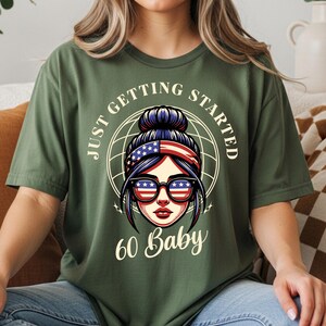 以下が含まれることがあります： オリーブグリーンのTシャツ。サングラスと愛国的なヘッドバンドをつけた女性のグラフィックが特徴です。Tシャツのテキストは「JUST GETTING STARTED」と「60 Baby」です。デザインは地球儀を背景にしています。