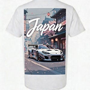 May include: White t-shirt with a graphic of a silver sports car on a Japanese street scene. The word "Japan" is written in a cursive font above the car. The background features traditional Japanese buildings and cherry blossoms.