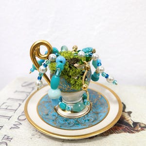 Puede incluir: Un juego de taza y platillo en miniatura con asa y ribete dorados. La taza está llena de musgo verde y adornada con cuentas turquesas, azules y perlas. El platillo tiene un diseño azul y dorado a juego. Una pieza decorativa caprichosa.