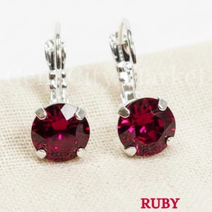 May include: A pair of ruby red leverback earrings. Each earring features a large, round, faceted ruby gemstone set in a silver-tone metal setting. The word "RUBY" is printed in red at the bottom of the image.
