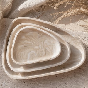 Könnte beinhalten: Ein Set aus drei ineinander stapelbaren, dreieckigen Schalen mit einem wirbelnden Marmormuster in Beige-, Creme- und Hellbrauntönen. Die Schalen haben glatte, abgerundete Kanten und ein flaches, offenes Innendesign, die ineinander liegen.