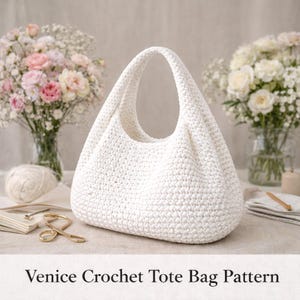 May include: A white, crocheted tote bag with a rounded shape and a single handle. The bag is made of a thick, textured yarn. The text "Venice Crochet Tote Bag Pattern" is at the bottom of the image. Flowers and crafting supplies are in the background.