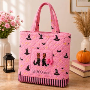 May include: A quilted pink tote bag featuring a pattern of black witch hats and bats. The front pocket displays a graphic of striped witch boots with a small pink bow and the text "fab-BOO-lous!" The base has black and pink vertical stripes.