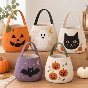 Puede incluir: Cinco cestas de tela para dulces de Halloween con asas. Una es naranja con cara de calabaza, una beige con un fantasma blanco, una beige con un gato negro, una morada con un murciélago negro y una beige con tres calabazas pequeñas.