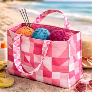 May include: A rectangular fabric tote bag featuring a pink and white geometric patchwork pattern with two matching handles. Inside the bag are three balls of yarn in orange, blue, and magenta, along with three silver knitting needles.